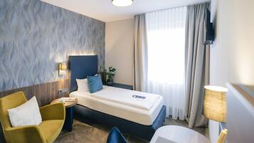 Una habitación de hotel con cama, un sillón amarillo, escritorio, lámpara y televisor.