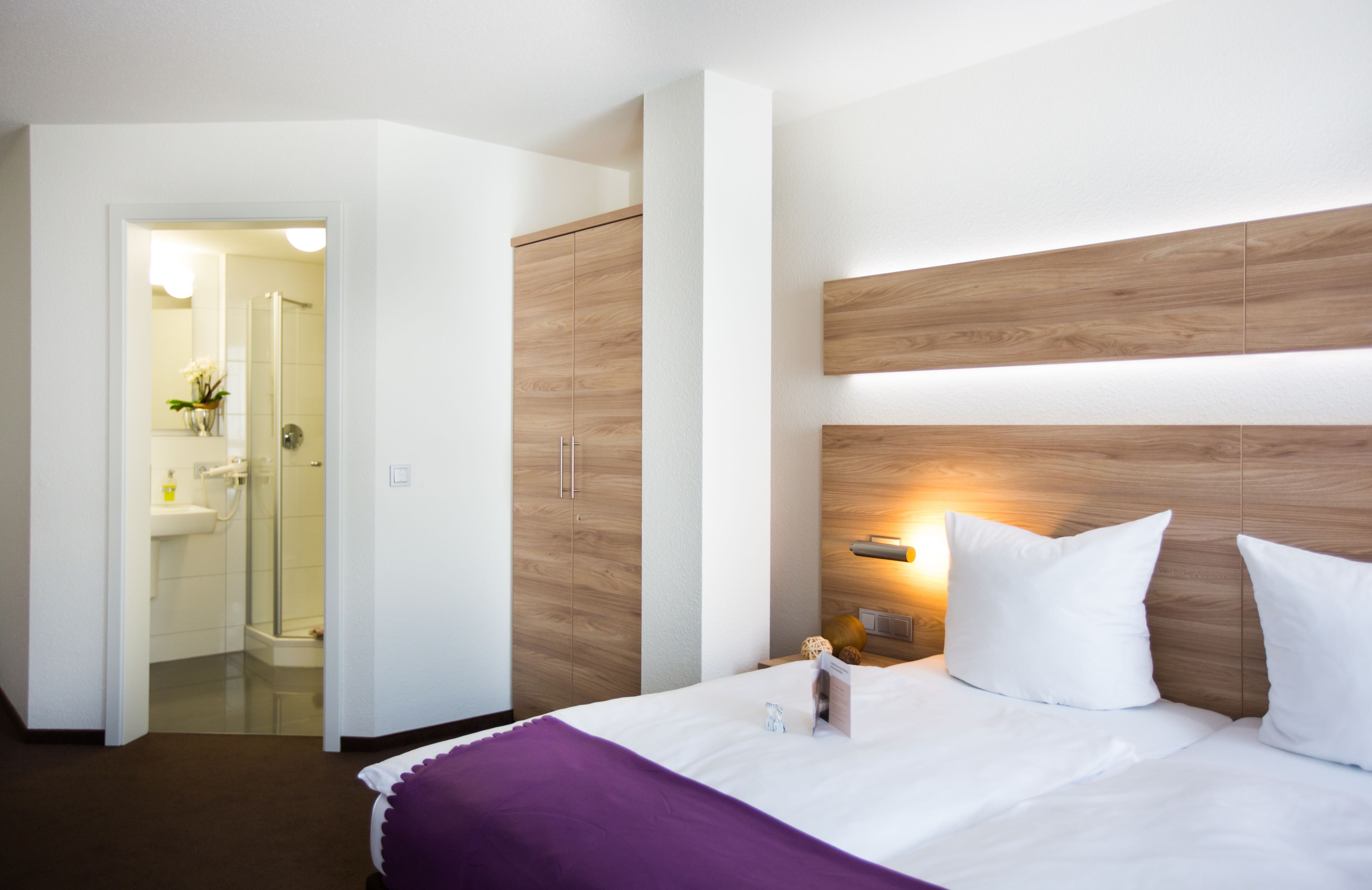 A hotel room with a large bed, a wooden headboard, a nightstand, and a bathroom visible through an open door.