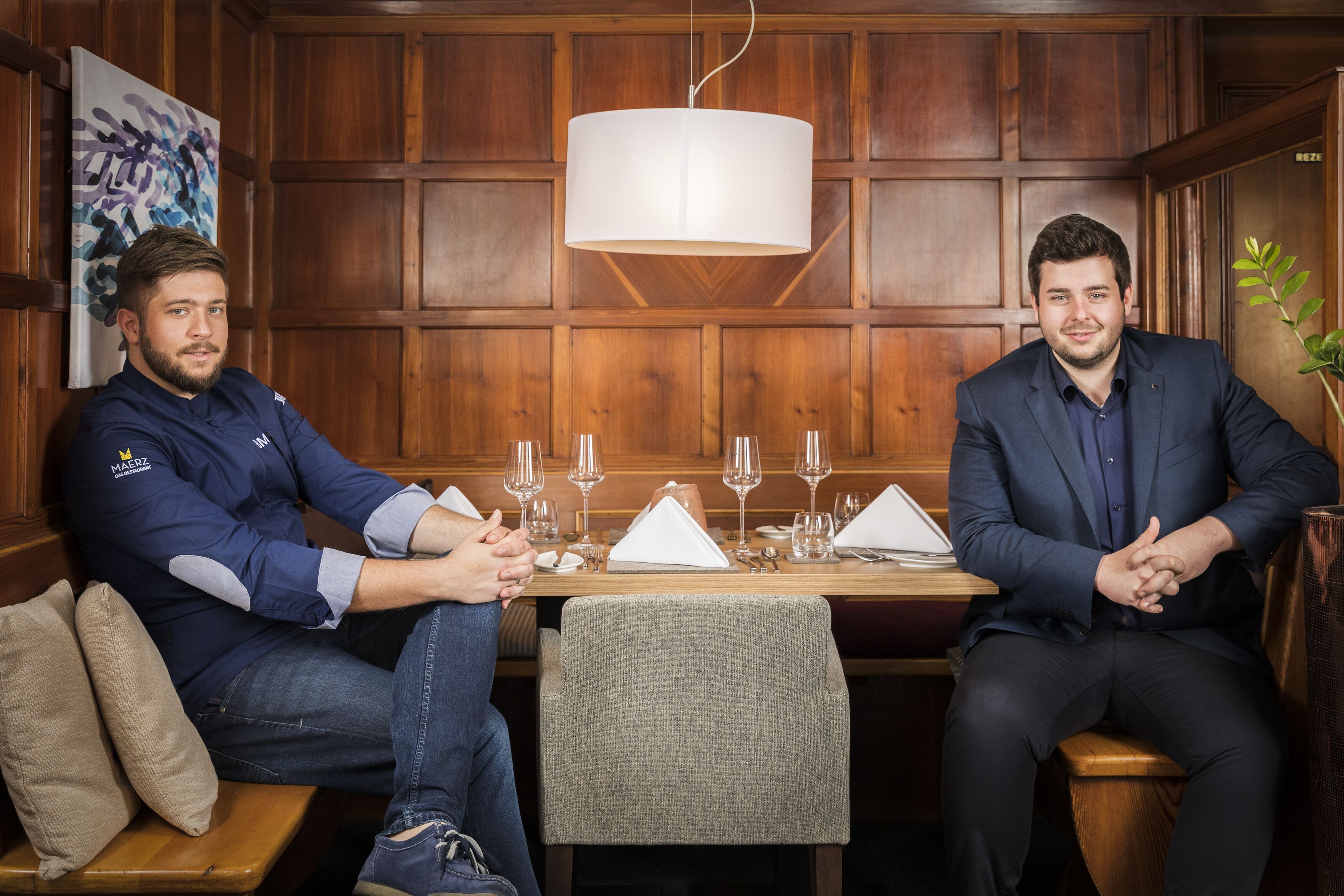 Two men seated at a wooden table with a white tablecloth, surrounded by wine glasses and a plant.