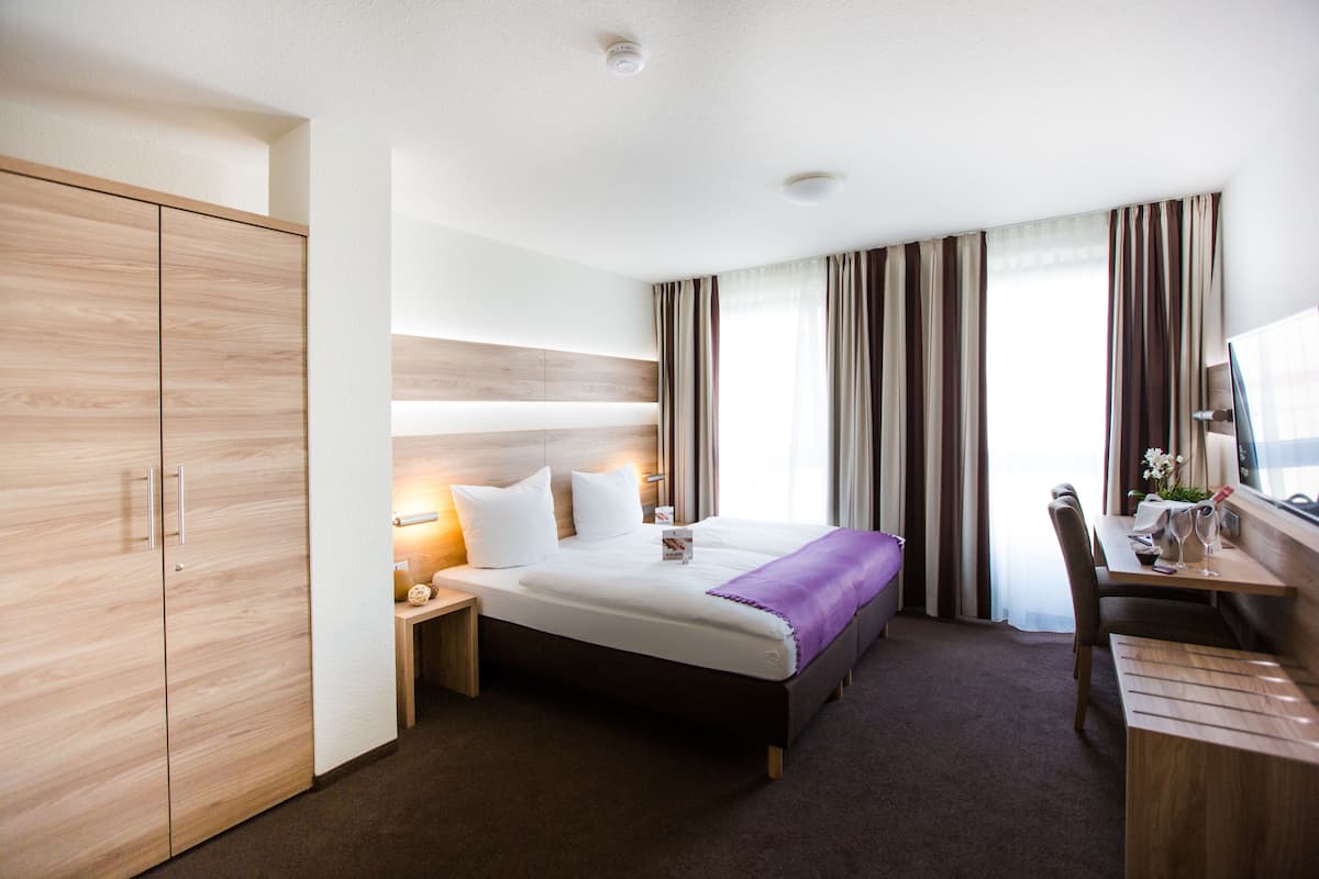 Ein Hotelzimmer mit Bett, Schreibtisch, Stuhl, Kleiderschrank und einem fensterbepflanzten Fenster.