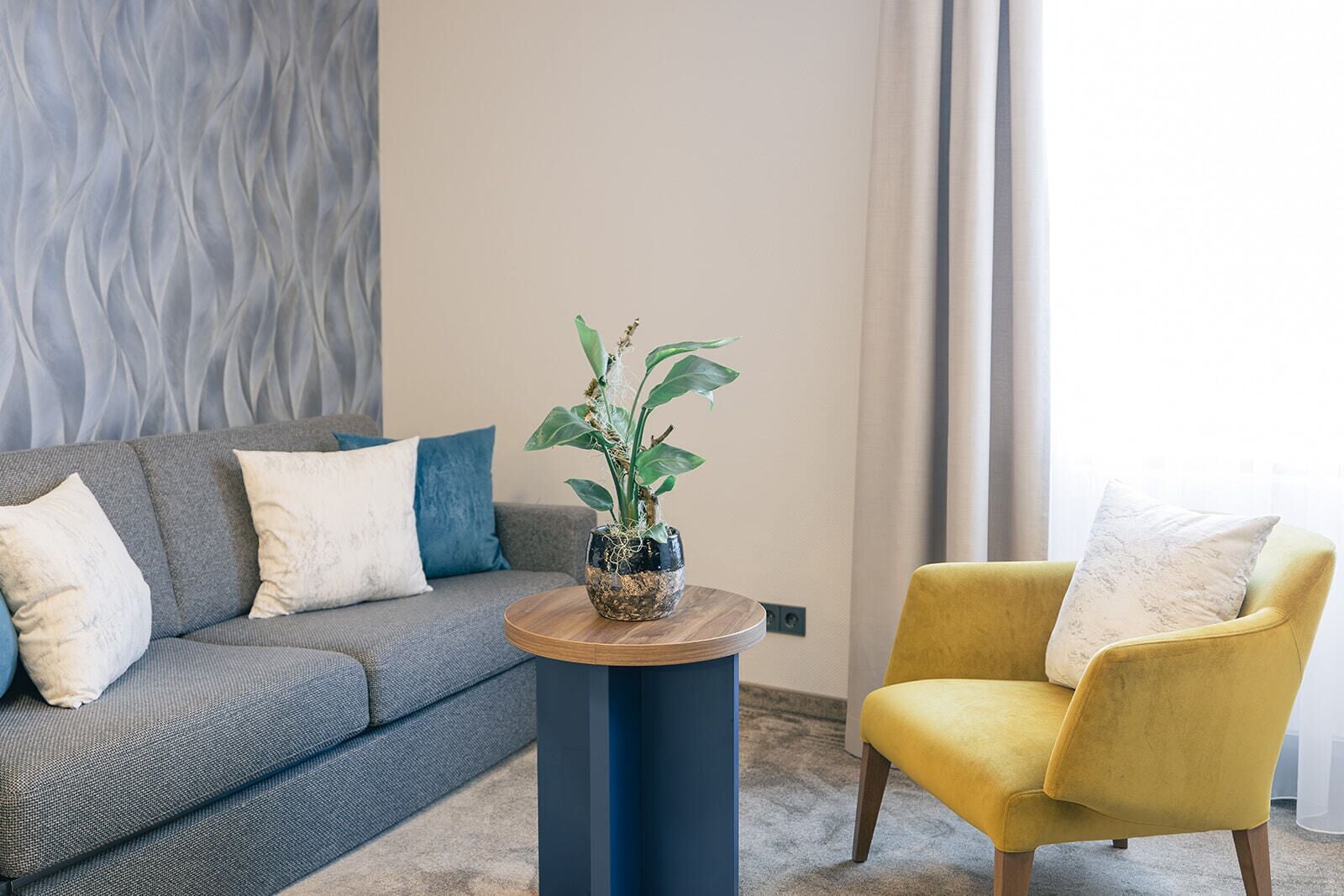 Una sala moderna con un sofá gris, un sillón amarillo y una mesa de centro redonda de madera con una planta en maceta.