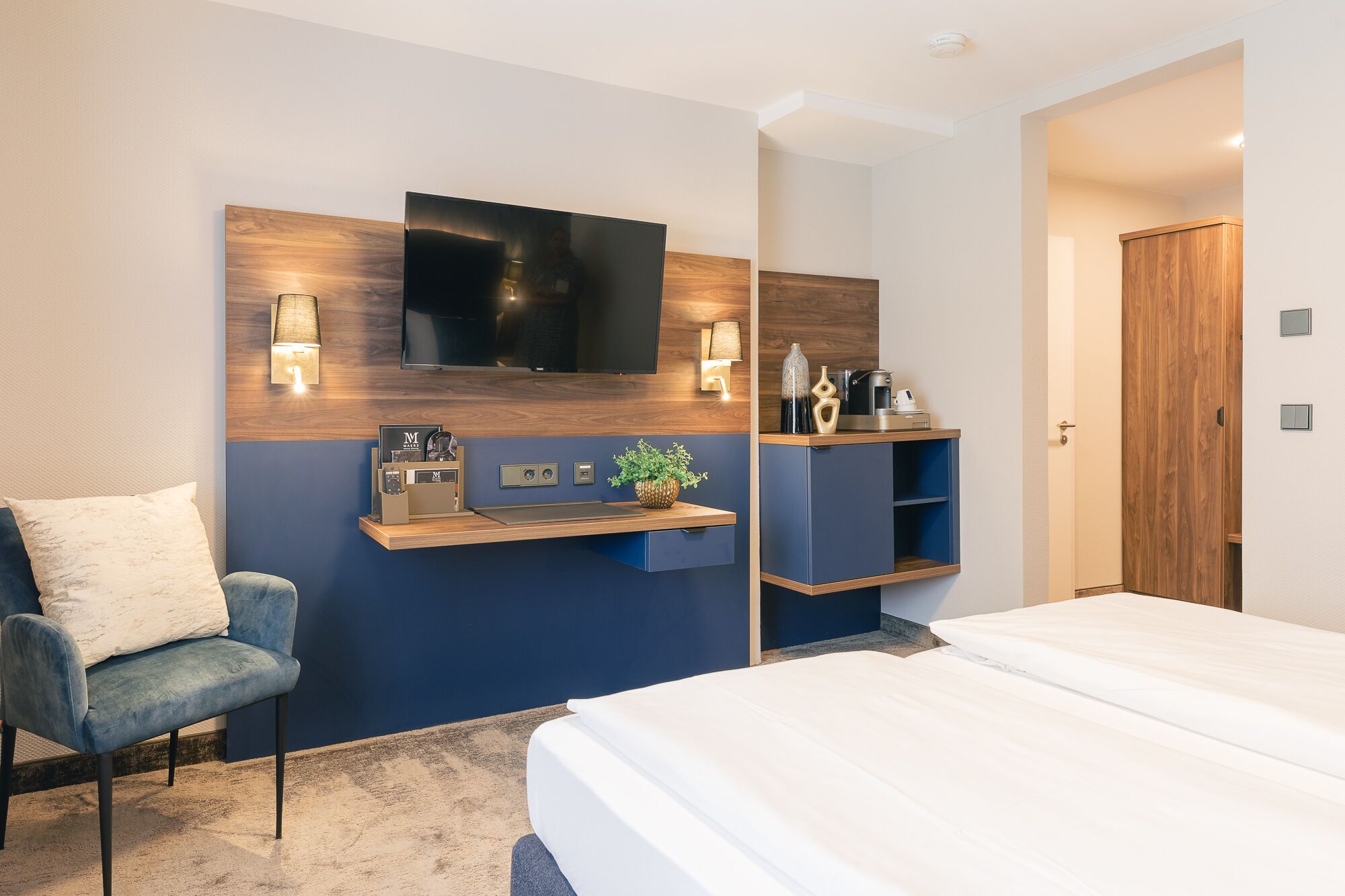 A modern hotel room with a bed, a desk with a flat-screen TV, a chair, and a wooden wardrobe.