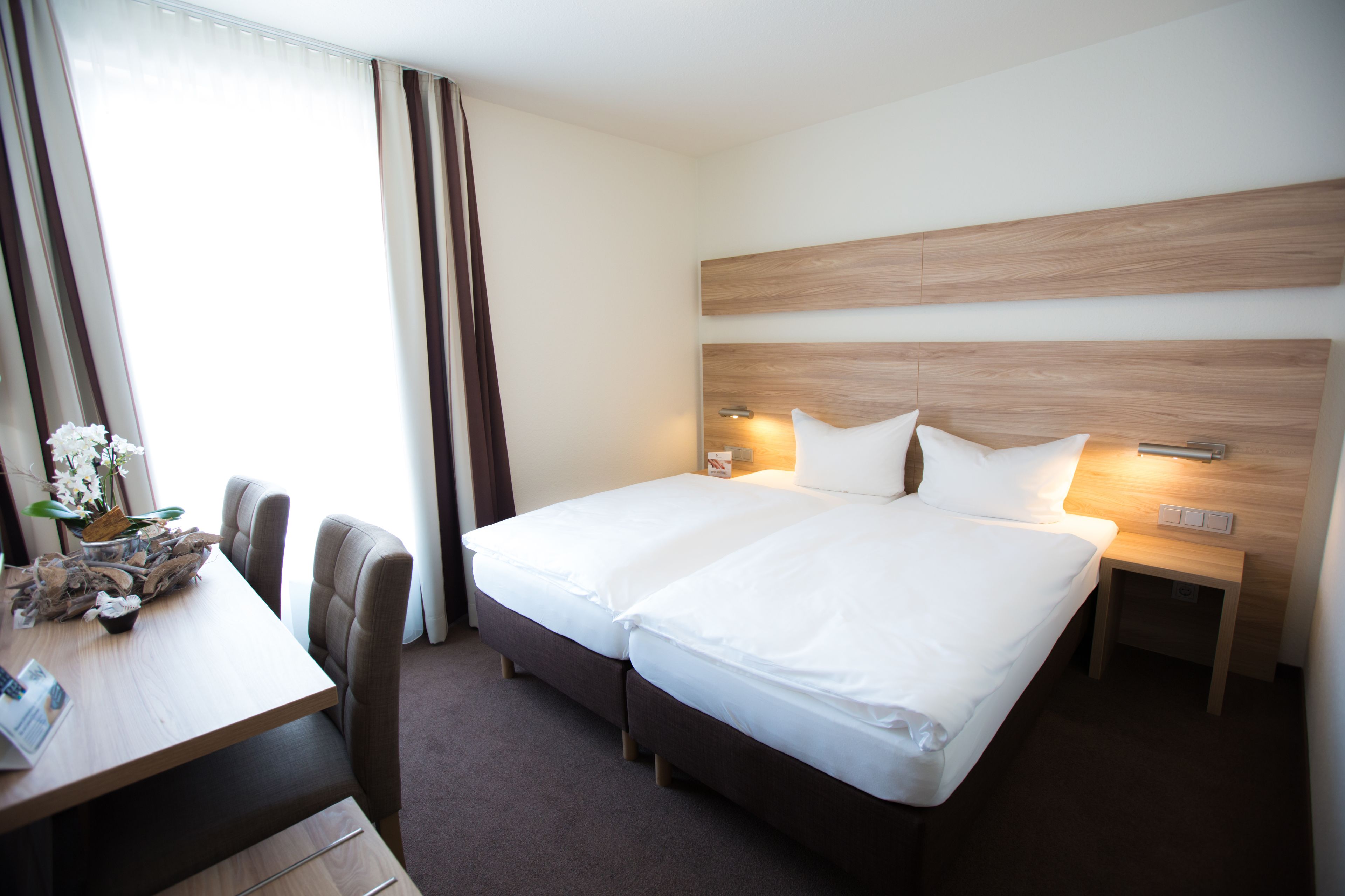 Une chambre d’hôtel avec un lit double, une tête de lit en bois, une table de chevet avec une lampe, une petite table sur laquelle est posé un vase de fleurs et une chaise.