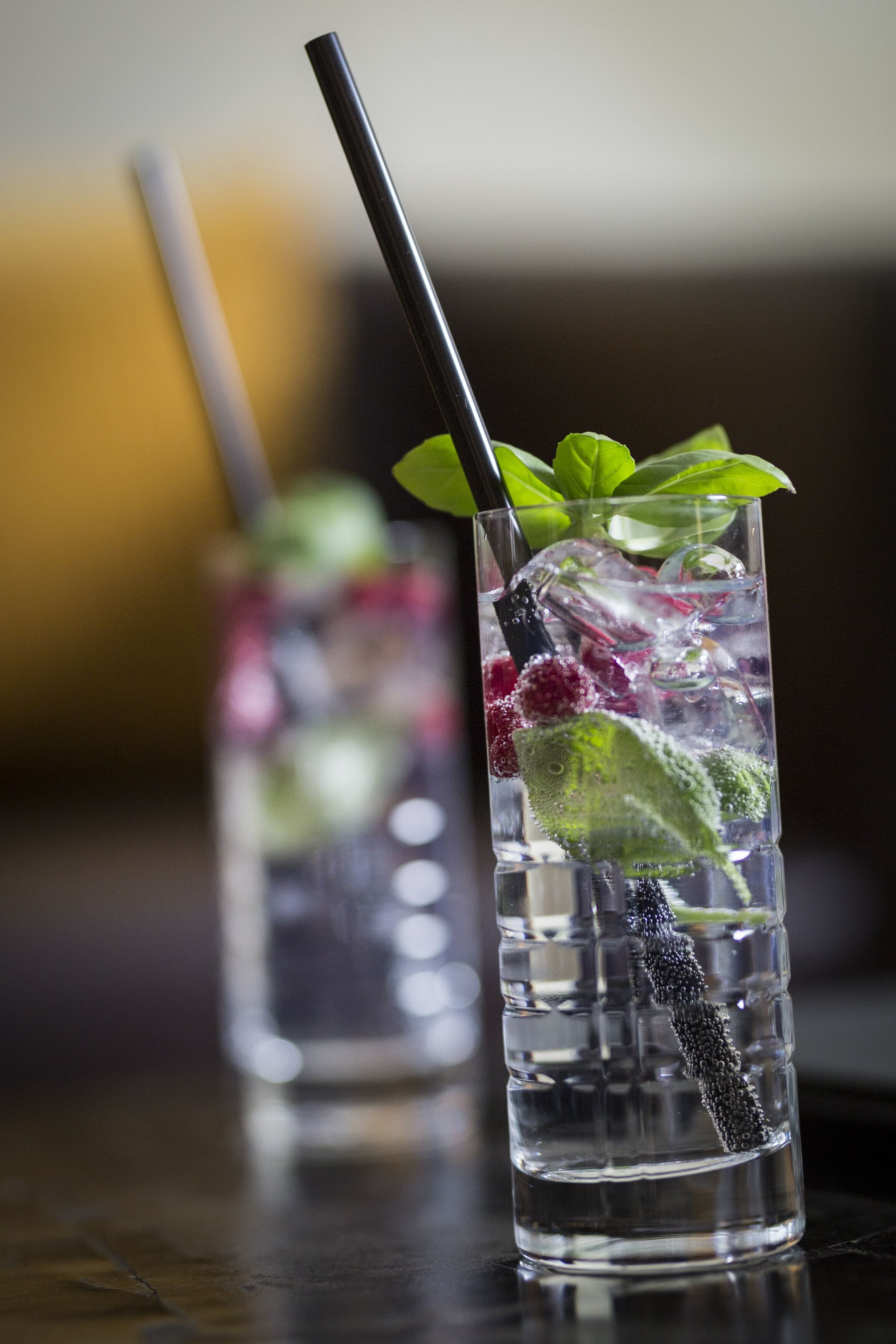 Two tall glasses of a clear beverage with ice, garnished with mint leaves and berries, and a black straw.