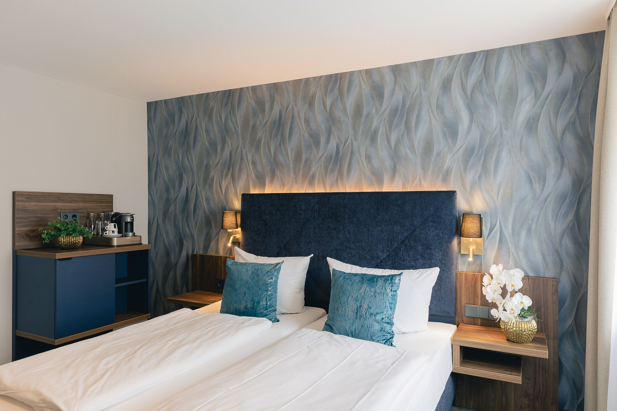 A modern hotel room with a large bed, two bedside tables with lamps, a small wooden desk, and a decorative plant.
