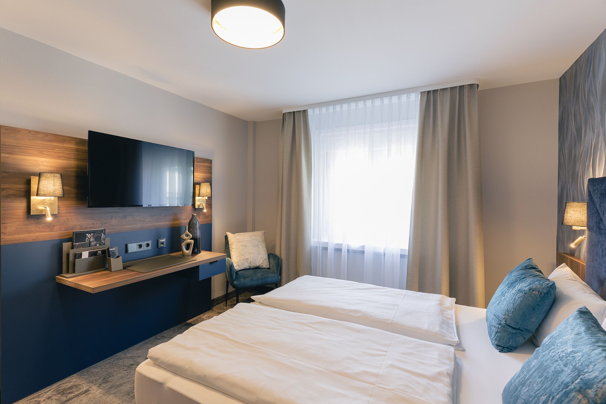A hotel room with a bed, a desk, a chair, a TV, and a window with curtains.