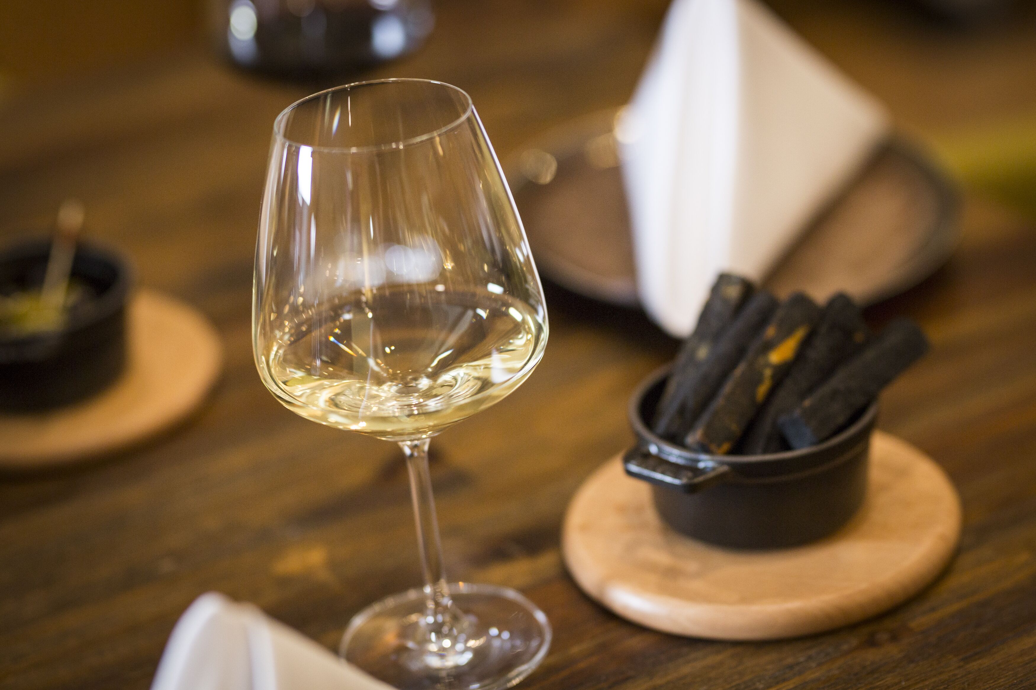 Una copa de vino sobre un posavasos de madera, con un cuenco pequeño de aceitunas y una servilleta a un lado.