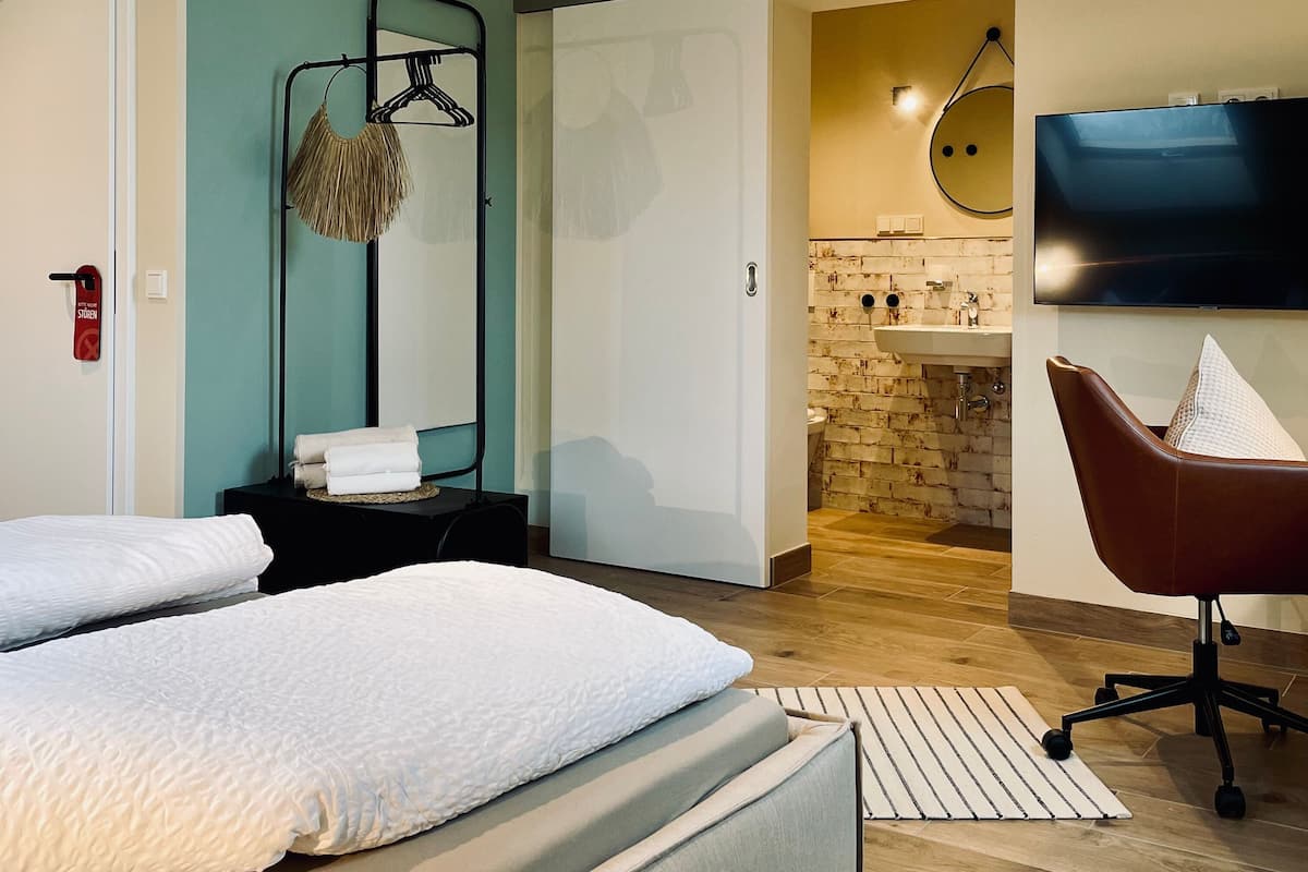 A modern hotel room with two beds, a sitting area with a chair, a TV, and a bathroom with a brick wall.