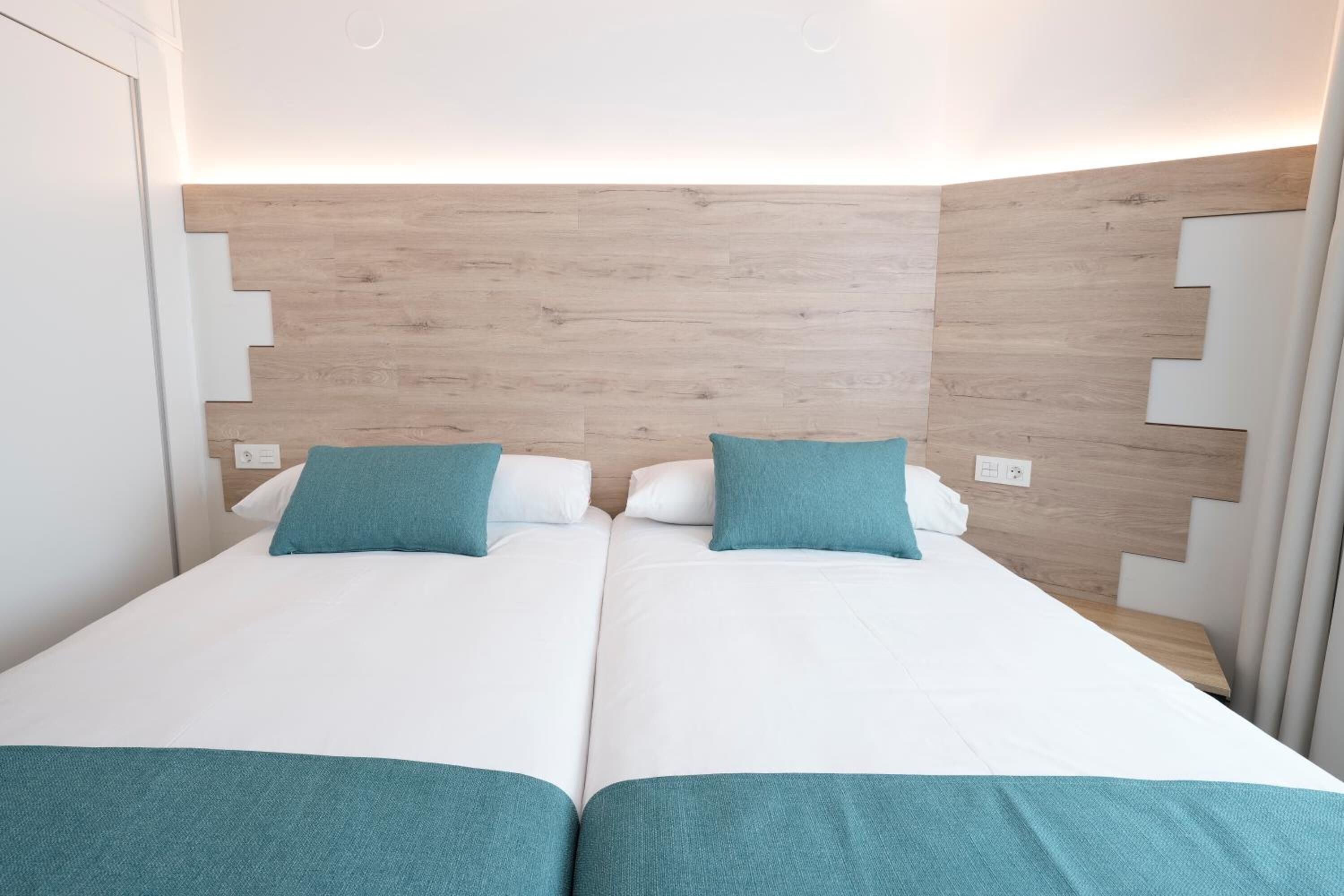 Ein Doppelbett mit weißer Bettwäsche und türkisfarbenen Kissen, vor einem Holzkopfende mit geometrischem Muster.