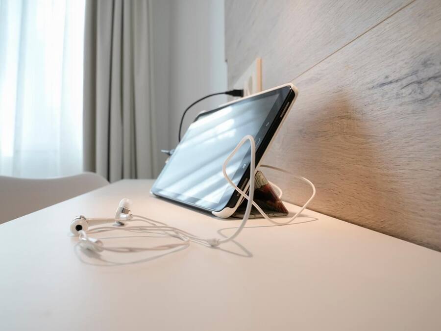 Una tableta sobre un soporte con auriculares sobre una mesa blanca en una habitación con cabecera de madera y una ventana con cortinas.