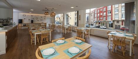 Un comedor amplio con mesas y sillas de madera, una cocina al fondo y ventanas grandes con vistas a la ciudad.