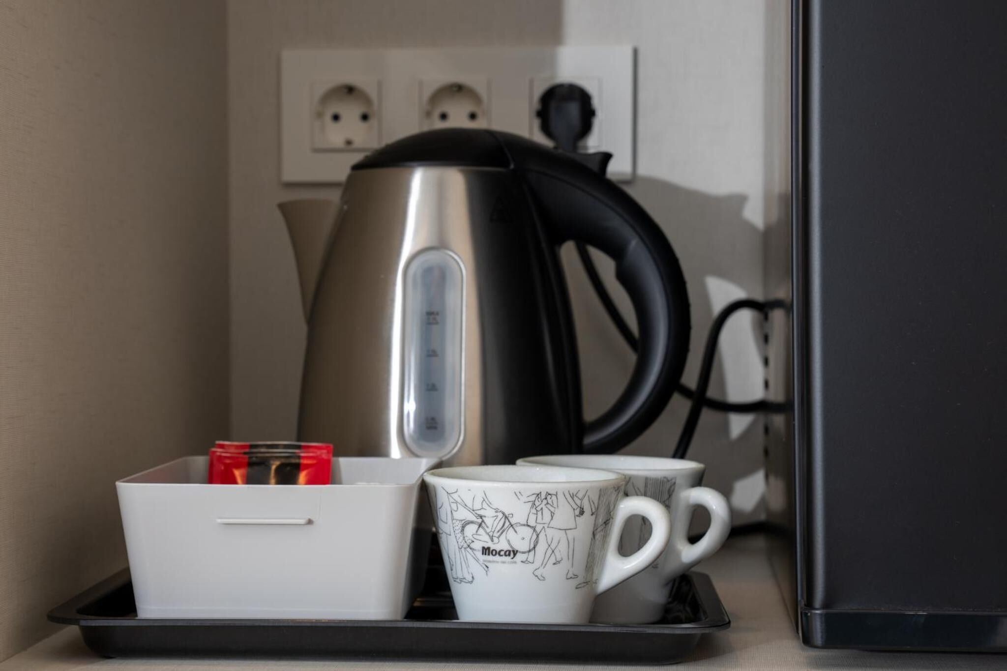 Een kitchenette met een waterkoker, een dienblad met mokken en een beker, en een stopcontact met drie aansluitingen.