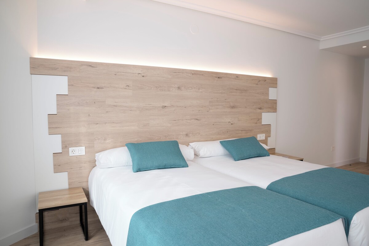 Una habitación de hotel moderna con cabecera de madera, dos camas con ropa de cama color turquesa, una mesita de noche pequeña y una mesita auxiliar.