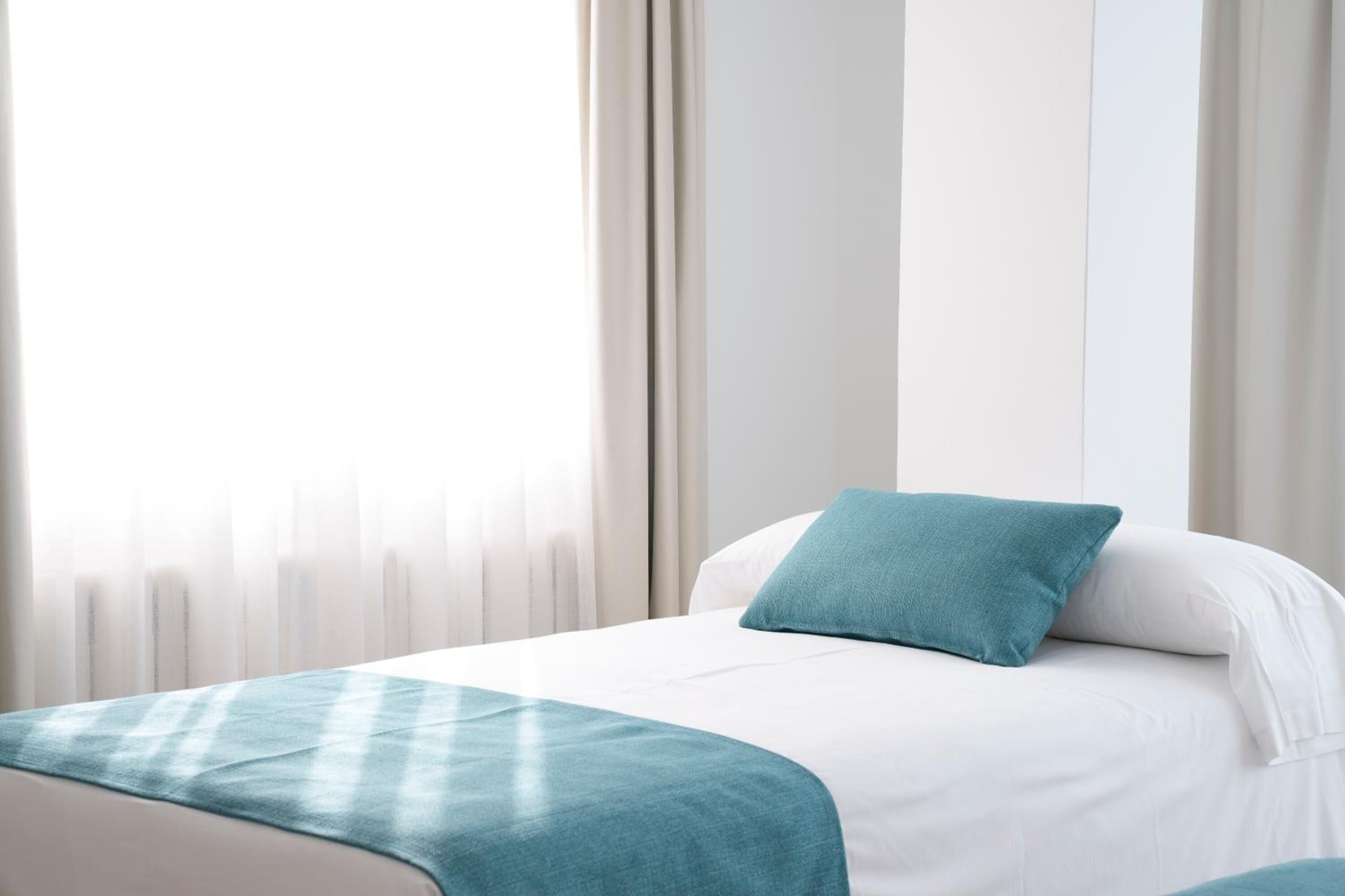 Ein Hotelzimmer mit einem Bett, einer petrolfarbenen Tagesdecke und transparenten Vorhängen.