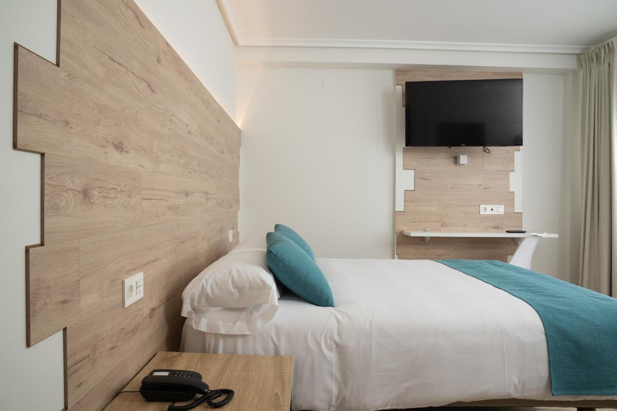 A hotel room with a wooden headboard, a bed with white bedding and teal pillows, a bedside table with a telephone, and a mounted flat-screen TV.