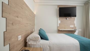 A hotel room with a wooden headboard, a bed with white bedding and teal pillows, a bedside table with a telephone, and a mounted flat-screen TV.
