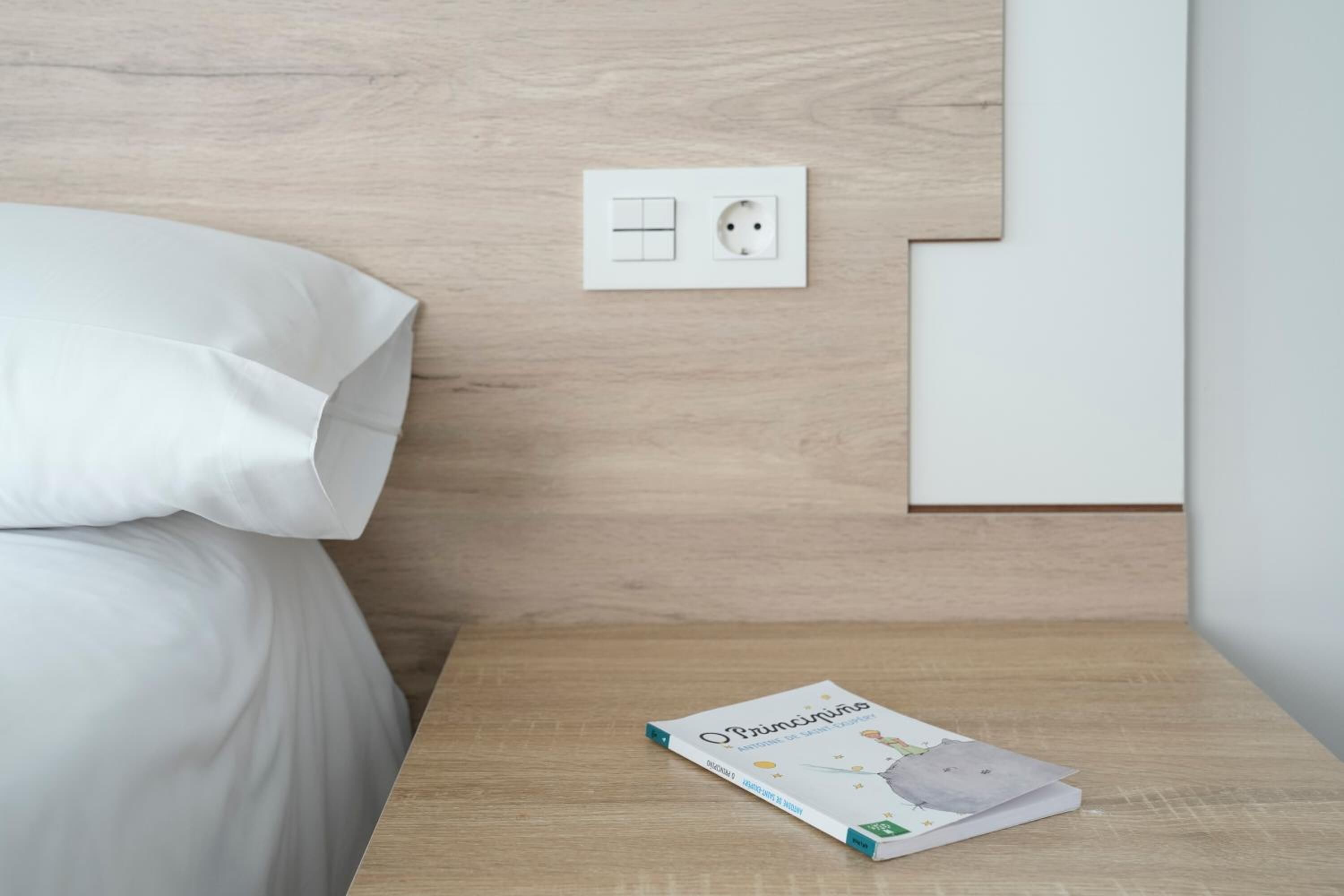 A neatly made bed with white bedding, a bedside table with a book, and a wall-mounted electrical outlet.