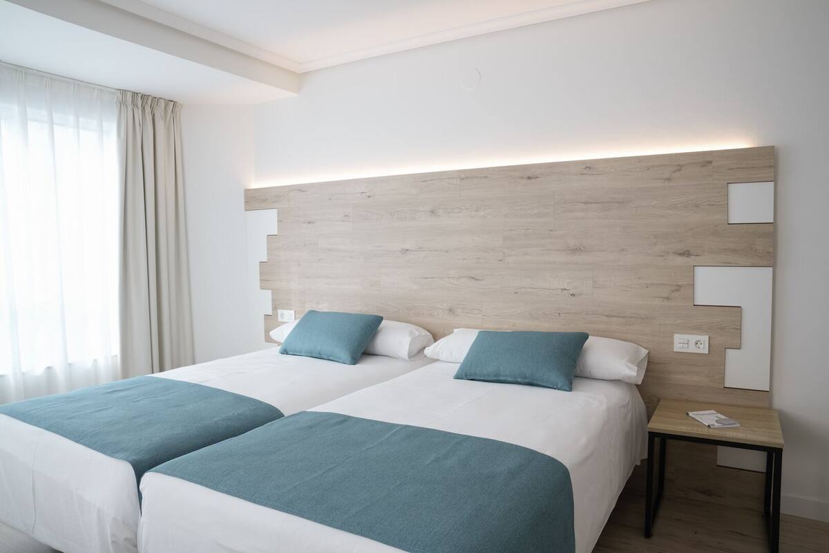 Une chambre d’hôtel avec deux lits, une tête de lit en bois, une table de chevet et une fenêtre avec des rideaux.