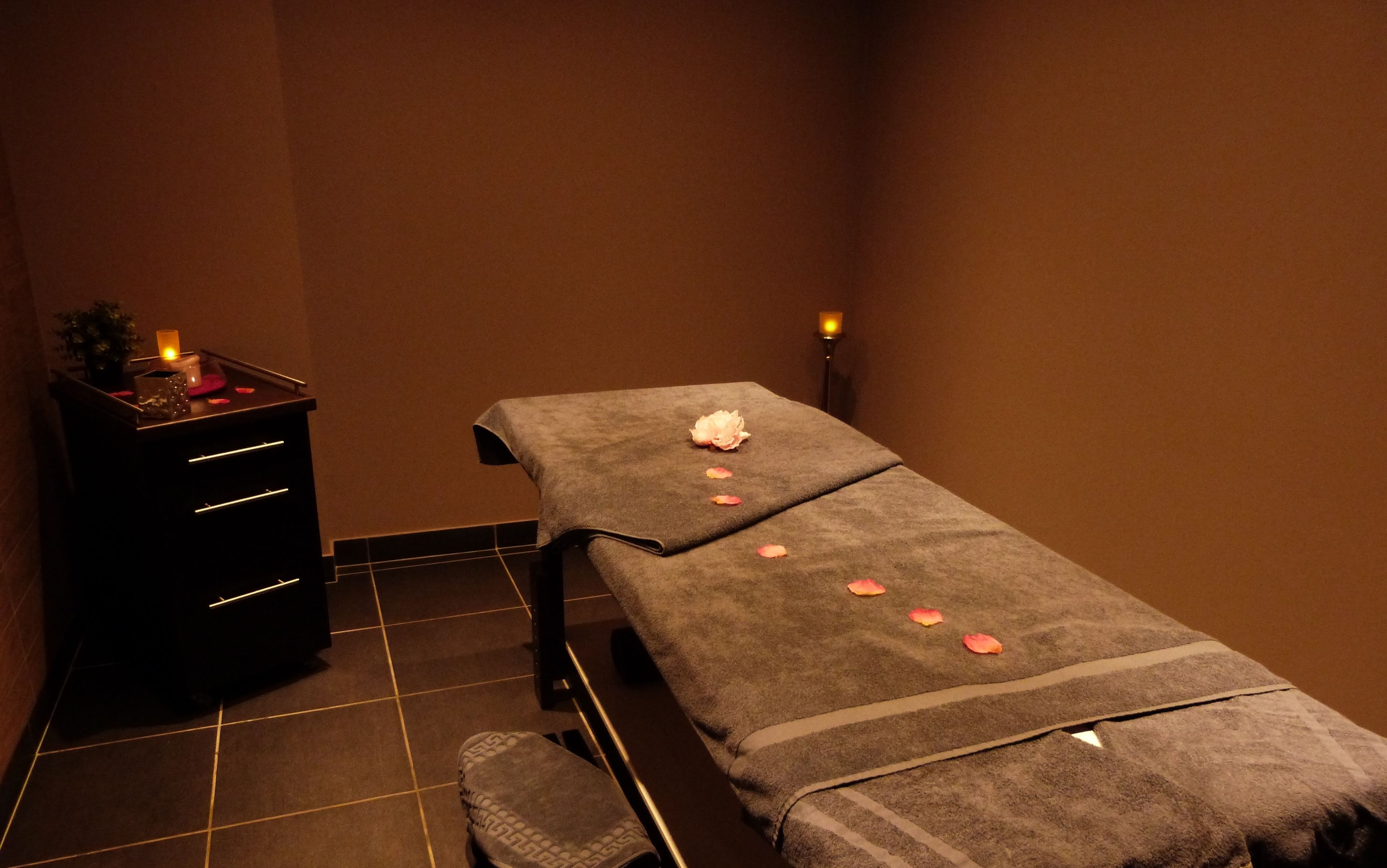 A massage table with a towel, rose petals, and a candle in a dimly lit room with a tiled floor and a small cabinet.