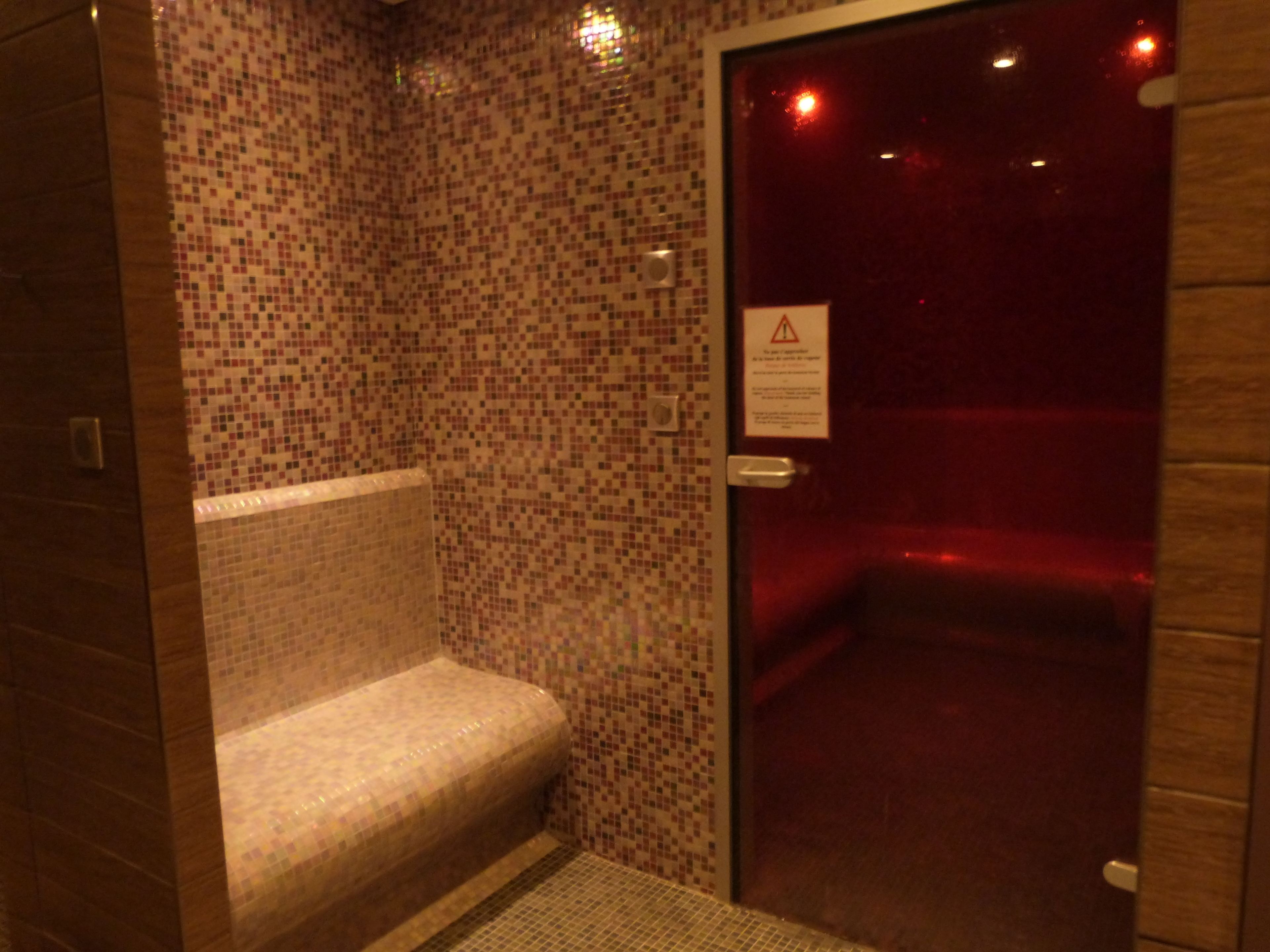 Un área de ducha con azulejos, un banco, una puerta con un letrero de advertencia y una pared revestida de azulejos.