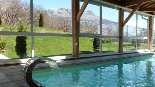 A swimming pool with a waterfall feature, large windows offering mountain views, and a wooden deck. - Ax'Hotel (Chorges)