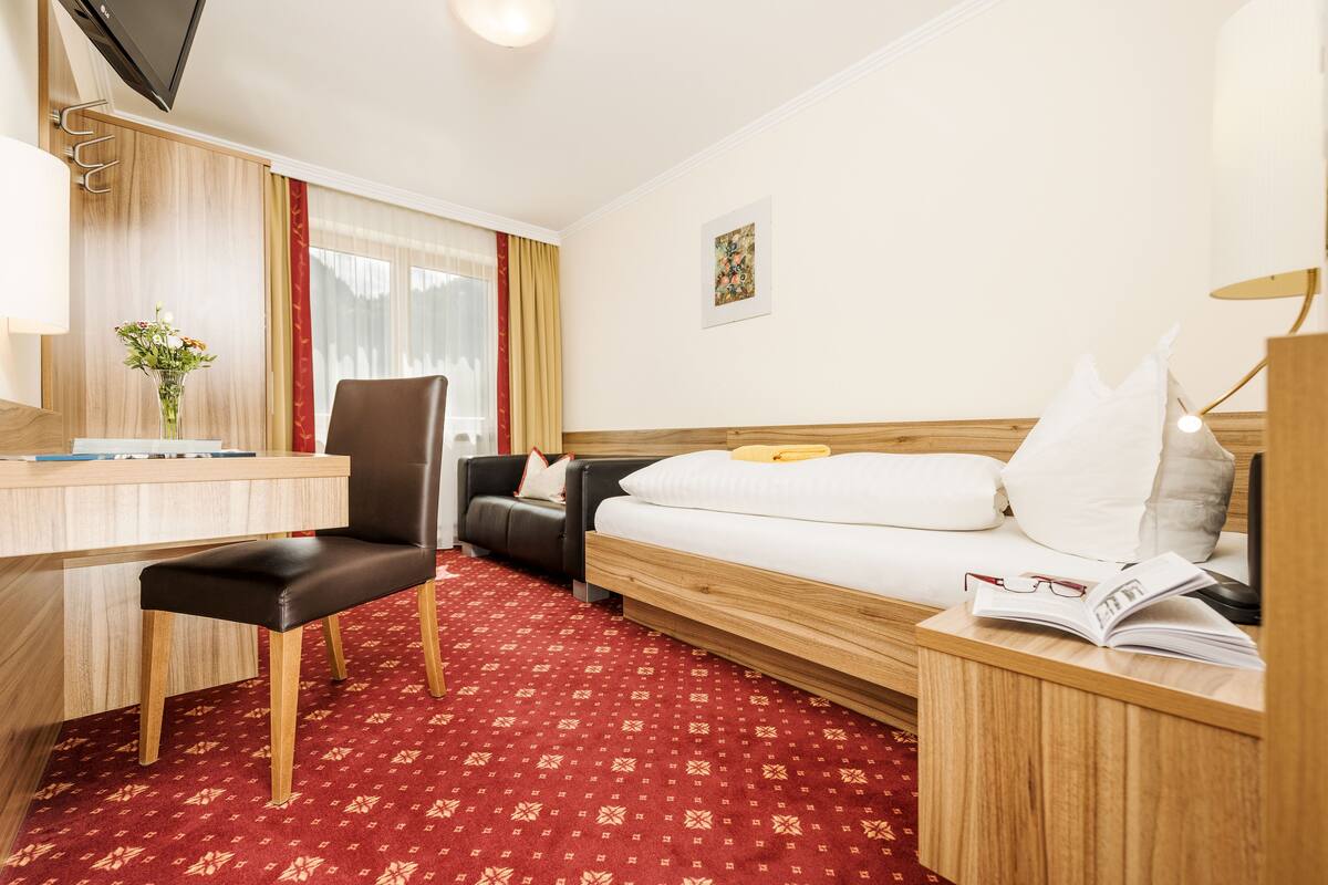 Habitación de hotel con cama, escritorio, silla, televisión, lámpara y ventana con cortinas.