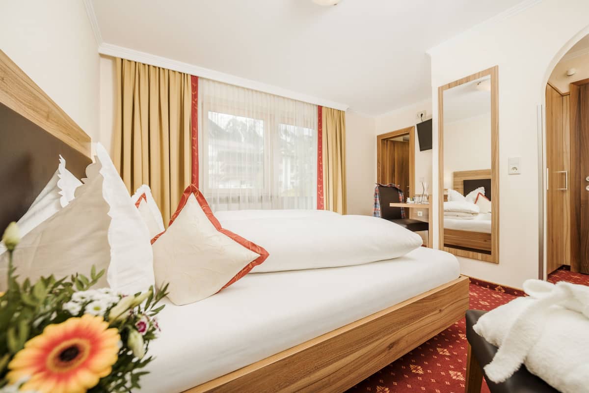 Habitación de hotel con cama, cabecero de madera, espejo grande, ventana con cortinas y pequeña mesa con lámpara.