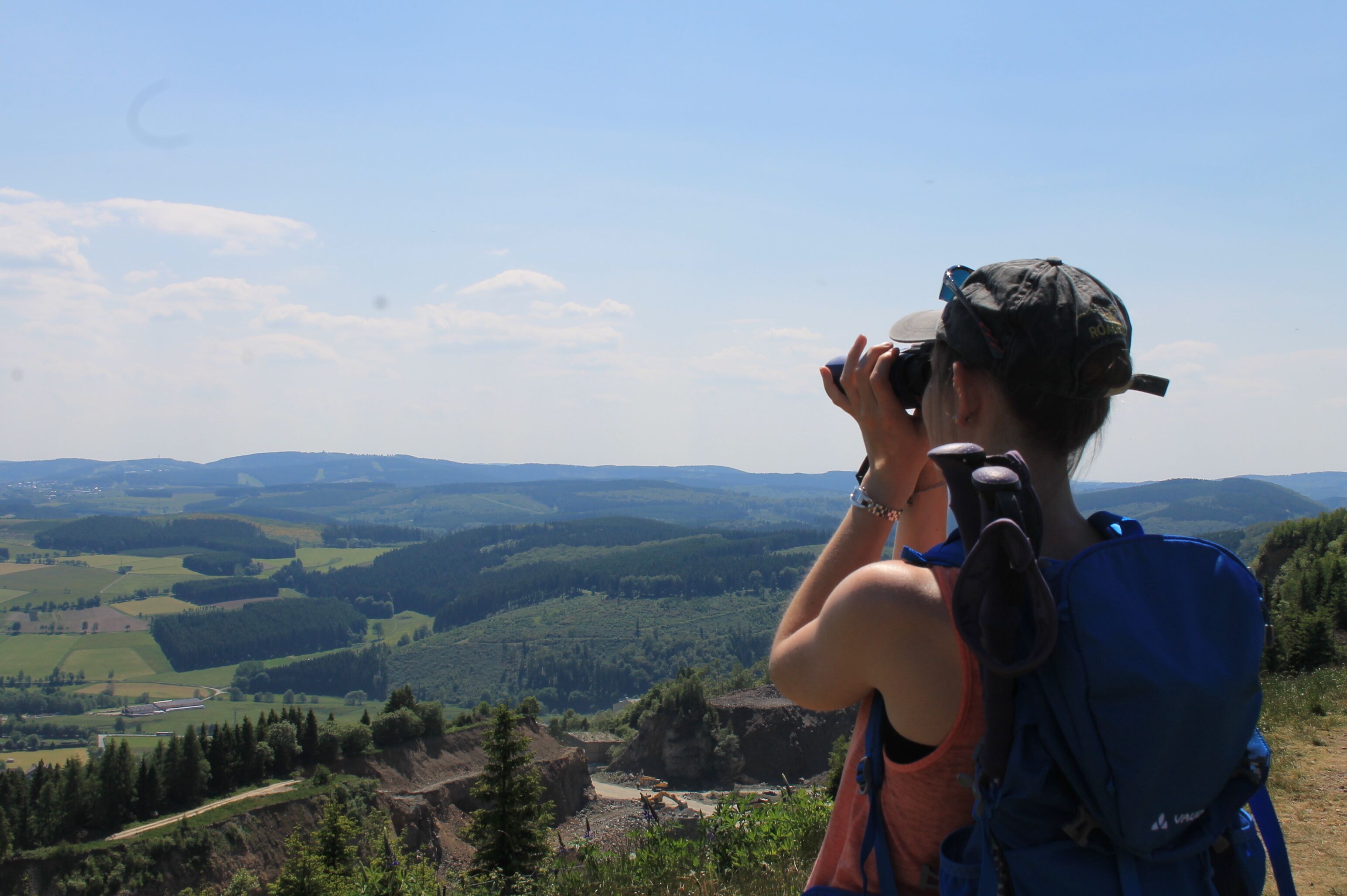 Iemand maakt een foto met een camera, draagt een pet en een rugzak en kijkt uit over een pittoresk landschap met heuvels en dalen.