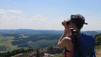 Iemand maakt een foto met een camera, draagt een pet en een rugzak en kijkt uit over een pittoresk landschap met heuvels en dalen.