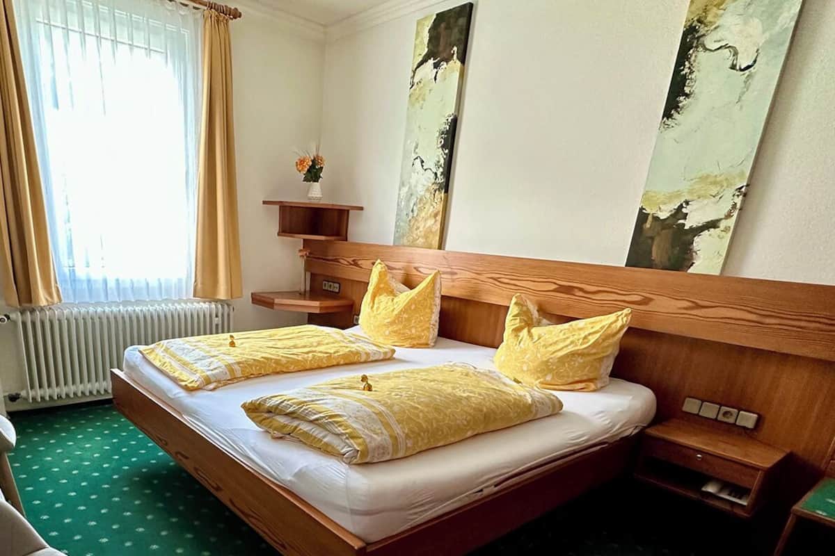 A hotel room with a double bed, two bedside tables, a chair, a desk with a lamp, a radiator, and artwork on the walls.