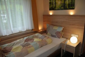 A bedroom with a bed, a bedside table with a lamp, a window with curtains, and a painting on the wall.