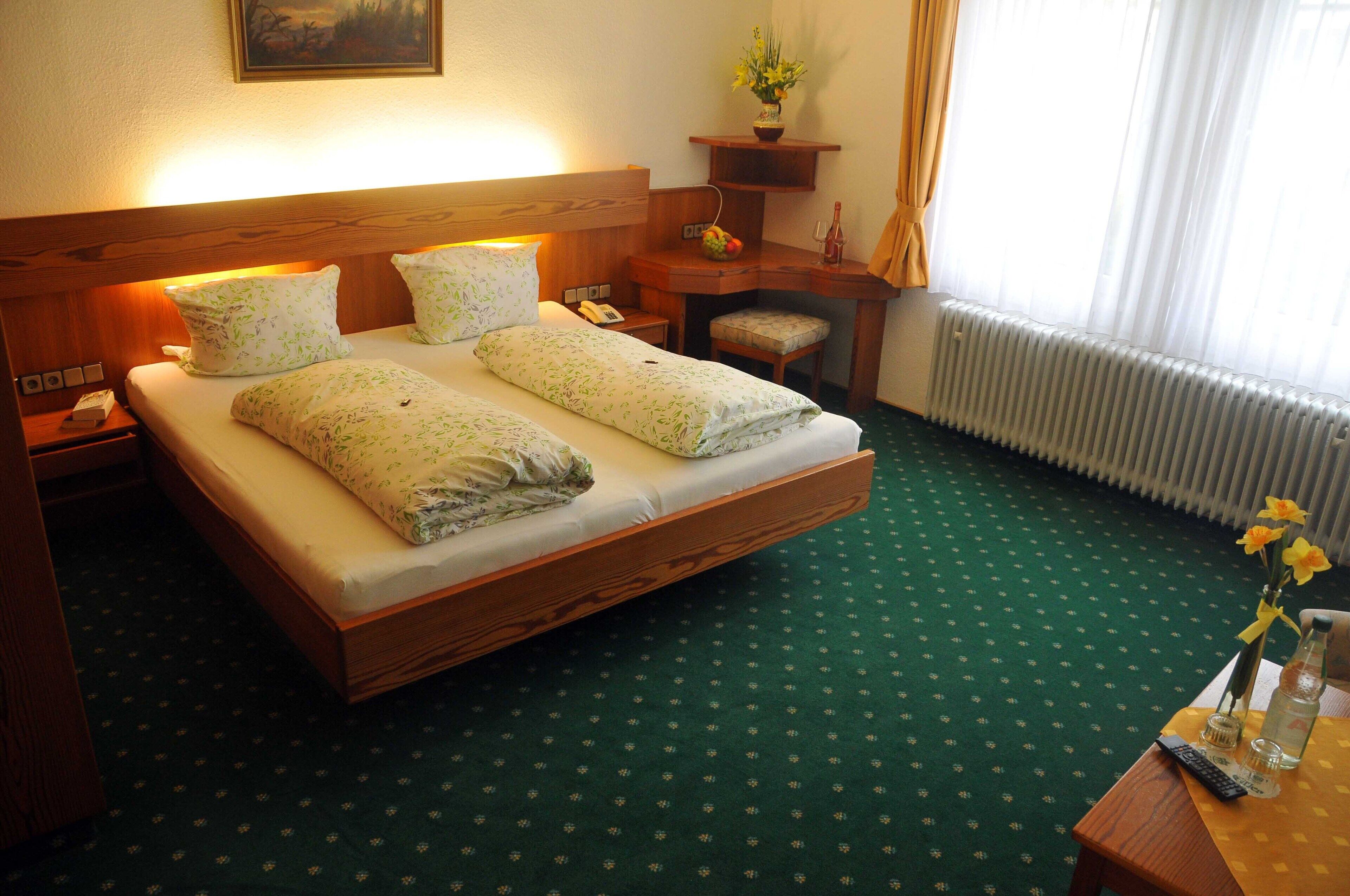 A double bed with floral bedding, a bedside table with a lamp, a chair, and a small table with a vase of flowers.