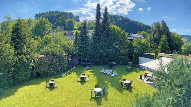 A lush garden with a variety of trees and shrubs, a grassy lawn with outdoor seating, and a view of a hill with buildings in the background. - Hotel Niedersfeld (Winterberg)