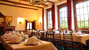 A dining area with multiple tables set for a meal, large windows with curtains, and a chandelier. - Hotel Niedersfeld (Winterberg)