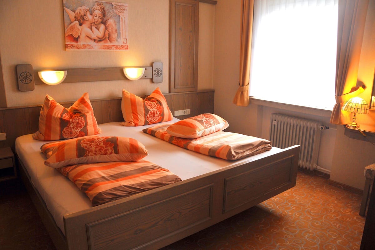A double bed with orange and brown striped bedding, a nightstand, a wall-mounted light, a radiator, and a window with curtains.