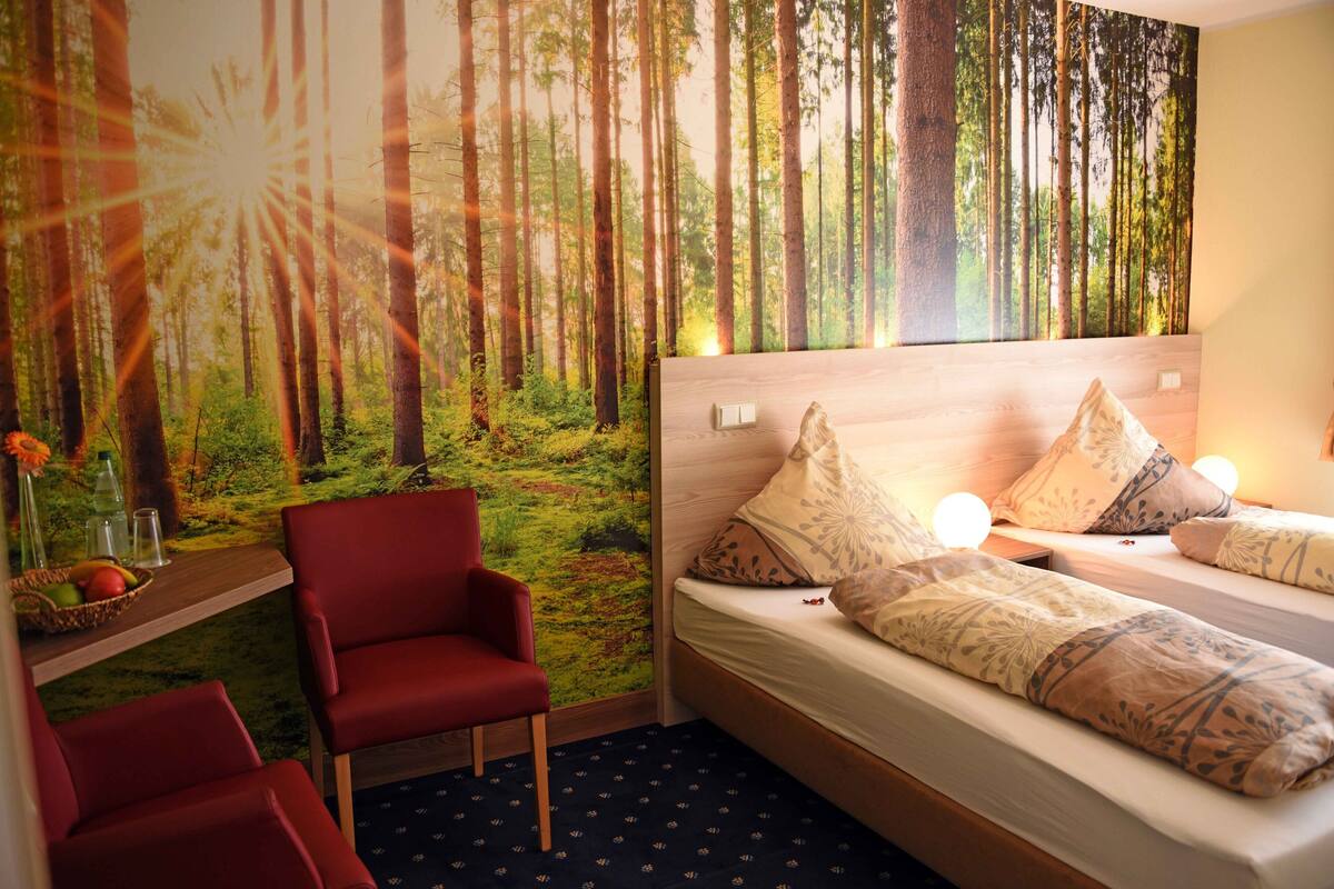 A room with a bed, red chairs, a shelf with fruits, and a forest mural on the wall.