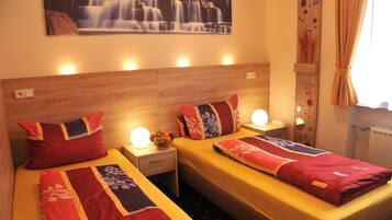 A hotel room with two beds, each with a red and blue patterned bedspread, a wooden headboard, bedside lamps, and a wall-mounted picture of a waterfall.