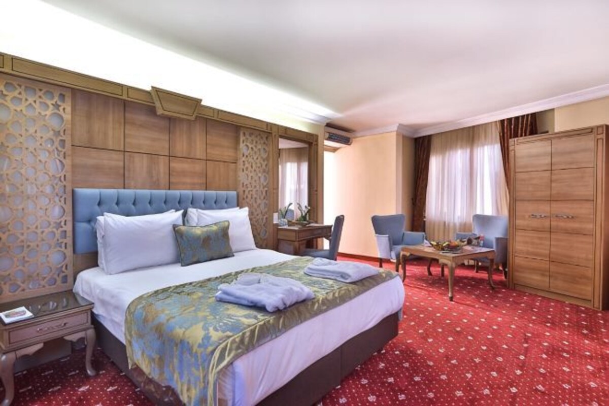 A hotel room with a large bed, a desk, two chairs, a wardrobe, and a carpeted floor.