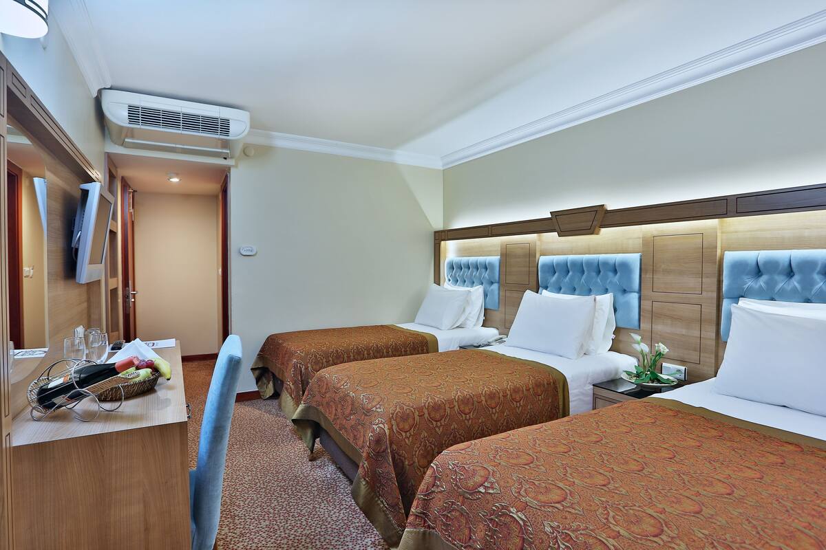A hotel room with two beds, a desk with a basket of fruit and a bottle, and a TV on the left.