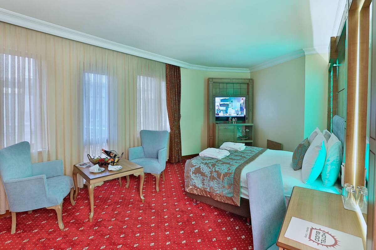 A hotel room with a red patterned carpet, a bed with a floral bedspread, a blue armchair, a wooden side table, and a television on a stand.