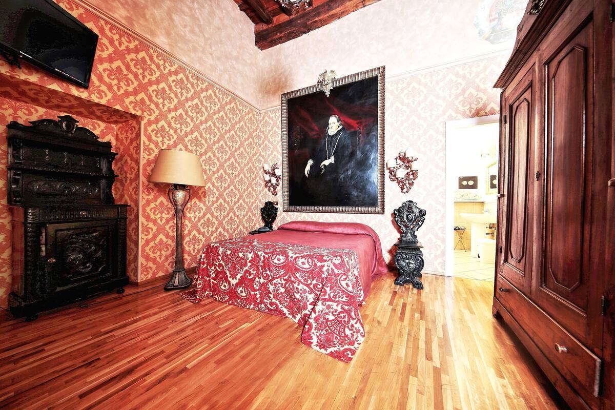 A bedroom with a wooden floor, a bed with a red and white patterned bedspread, a dark wooden dresser, a wall-mounted television, and a large framed portrait.