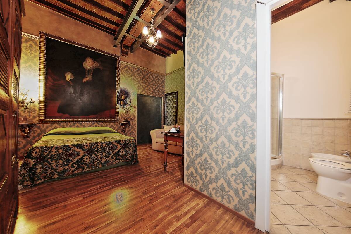 A room with a bed, a desk, a chair, a large painting, and a bathroom with a toilet and shower.