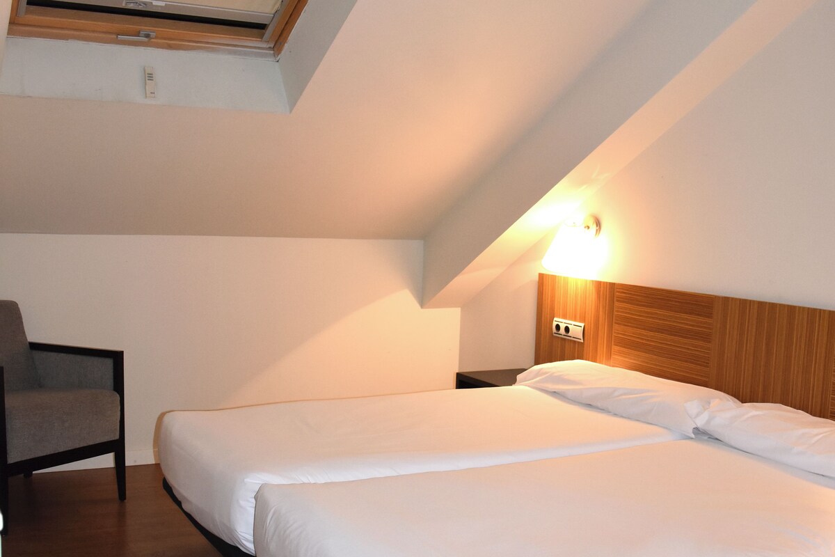 Un dormitorio con dos camas, una silla, una mesita de noche con lámpara y un tragaluz.