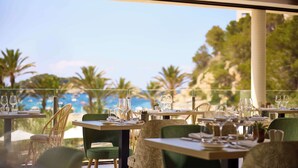 A restaurant with tables set for dining, overlooking a beach with palm trees and a clear sky.