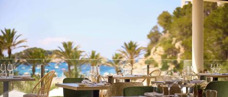 A restaurant with tables set for dining, overlooking a beach with palm trees and a clear sky.