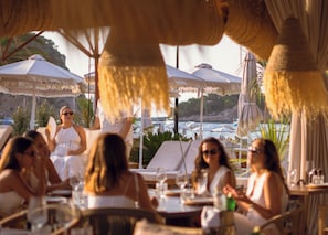 Un groupe de personnes est assis à la terrasse d’un restaurant, à l’ombre de parasols, et se détend sur des chaises longues.
