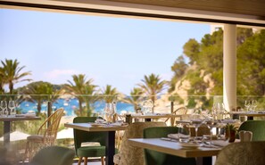 A restaurant with tables set for dining, overlooking a beach with palm trees and a clear sky. - Cala San Miguel Ibiza Resort, Curio Collection by Hilton (Sant Joan de Labritja)