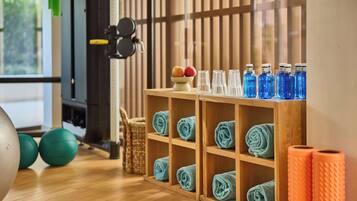 Un área de gimnasio con equipo de ejercicio, un estante de madera con toallas y botellas de agua, y una ventana con persianas.