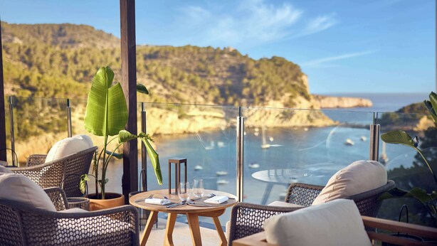 A balcony with wicker furniture, a round table with glasses and cutlery, and a view of a coastal landscape with cliffs and the sea. - Cala San Miguel Ibiza Resort, Curio Collection by Hilton (Sant Joan de Labritja)