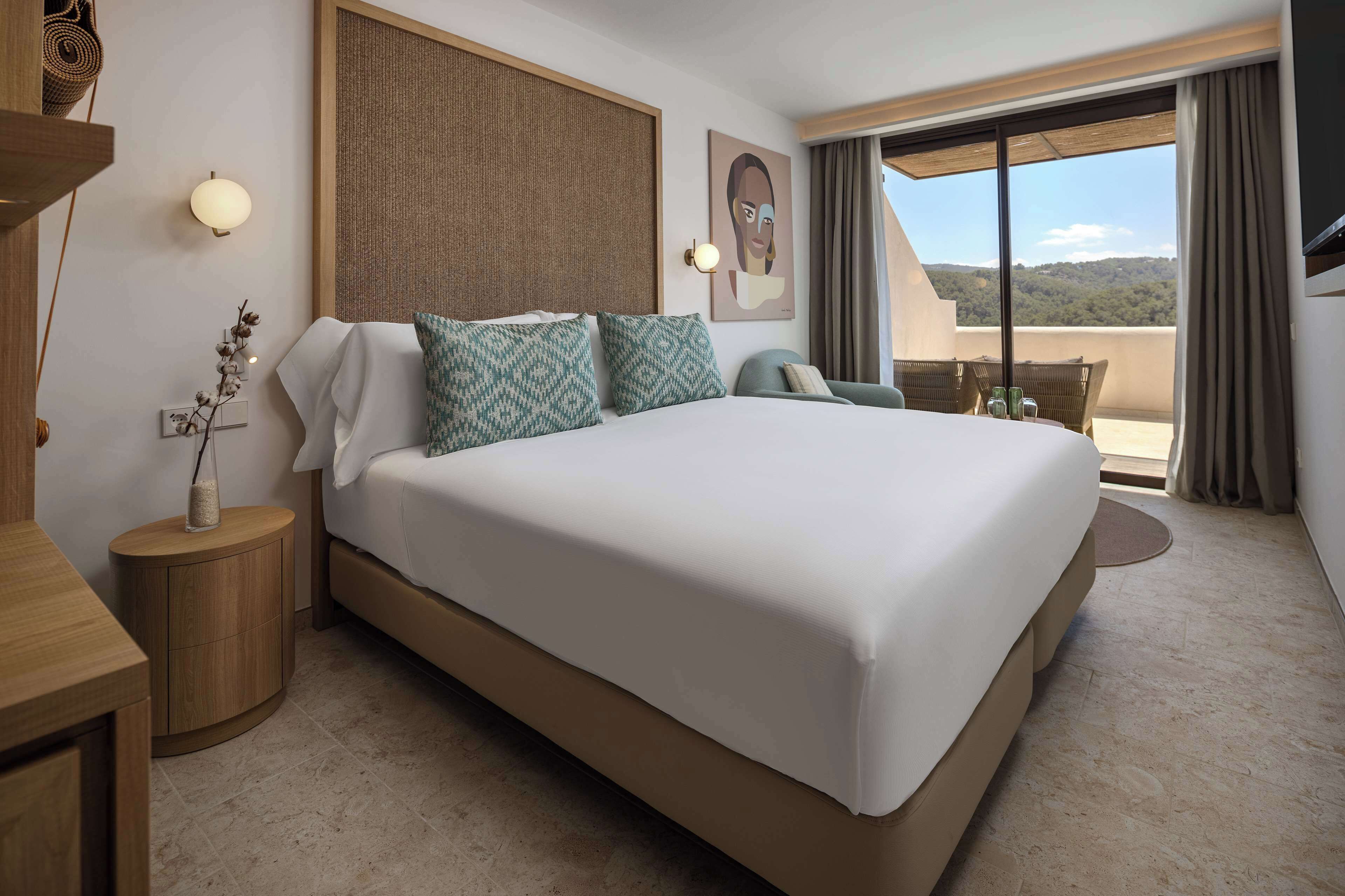 A hotel room with a large bed, a small side table, a wall-mounted lamp, a painting, and a balcony with a view.